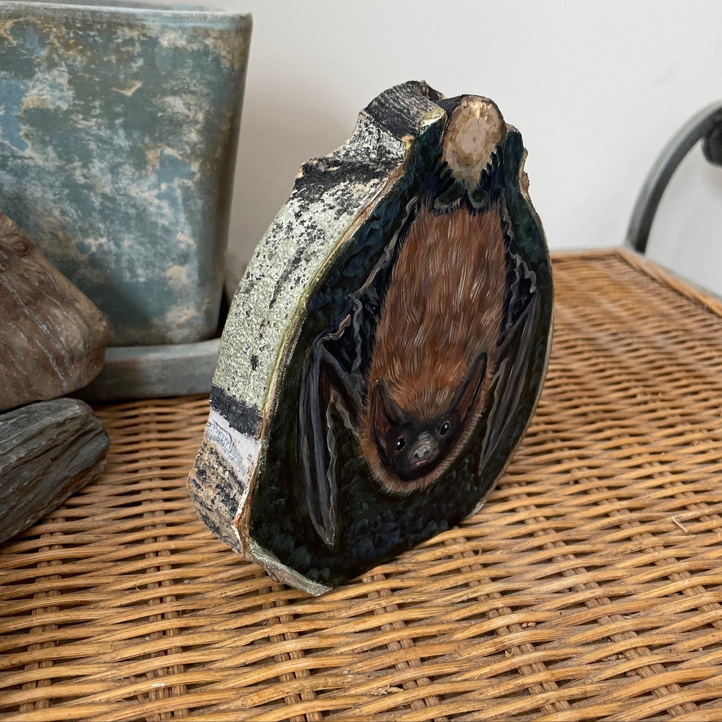 Painting of a Brown bat hanging upside down from a tree knot against a dark forest green background on an aspen wood slice with bark framing the outside