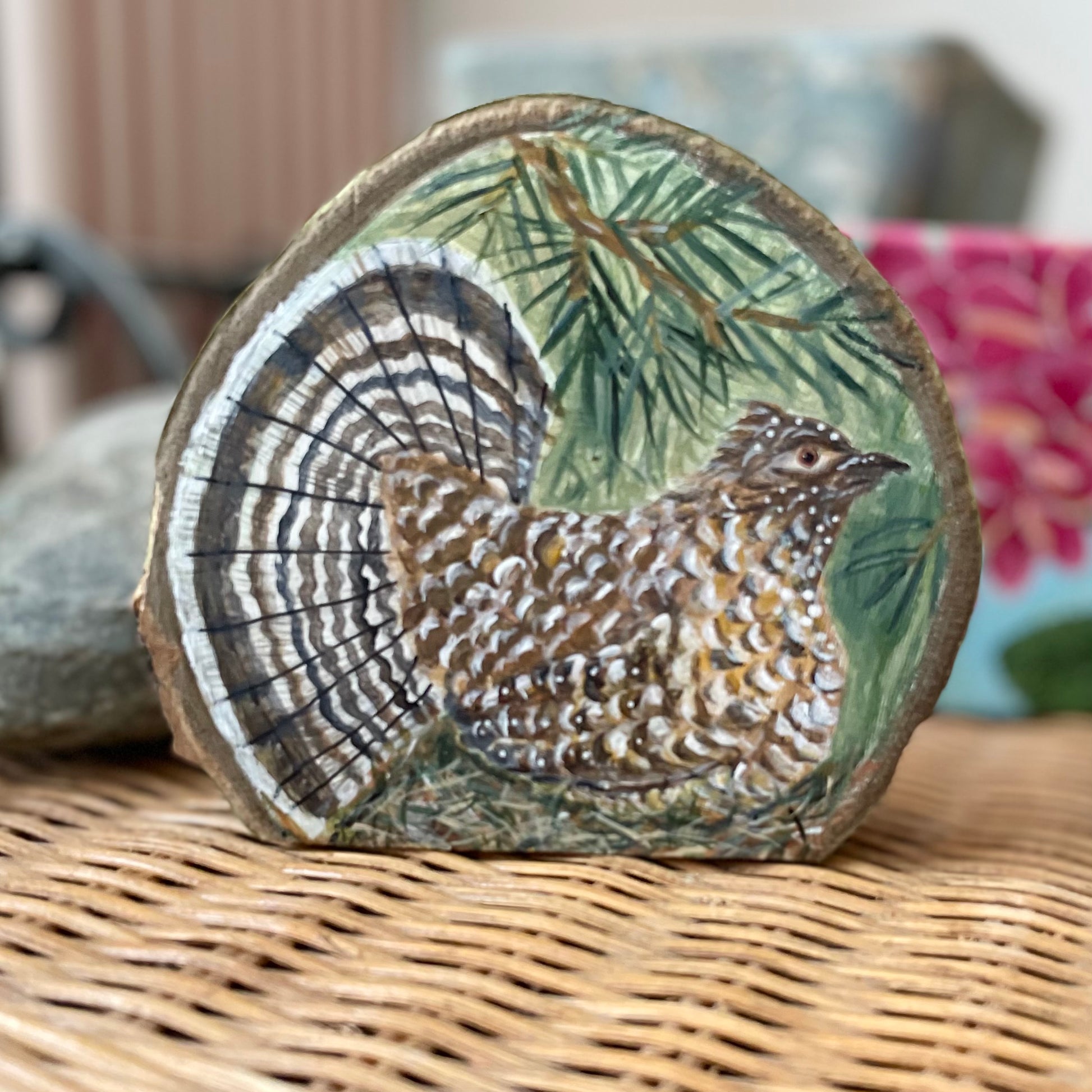 Painting of a ruffed grouse with open tail feathers under a pine branch on aspen wood slice with bark framing the outer edges 