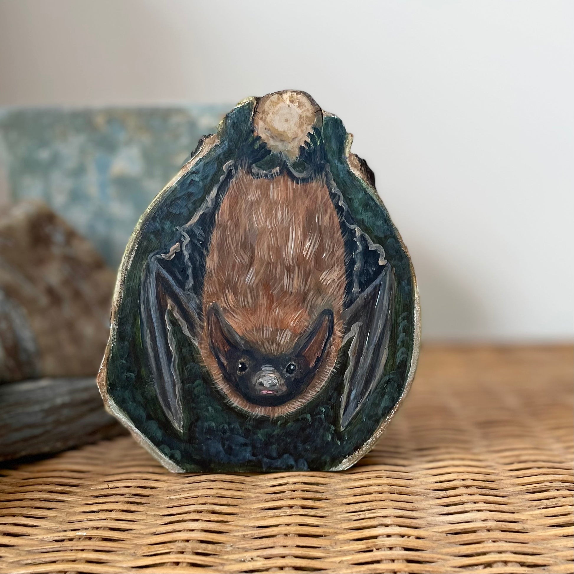 Painting of a Brown bat hanging upside down from a tree knot against a dark forest green background on an aspen wood slice with bark framing the outside