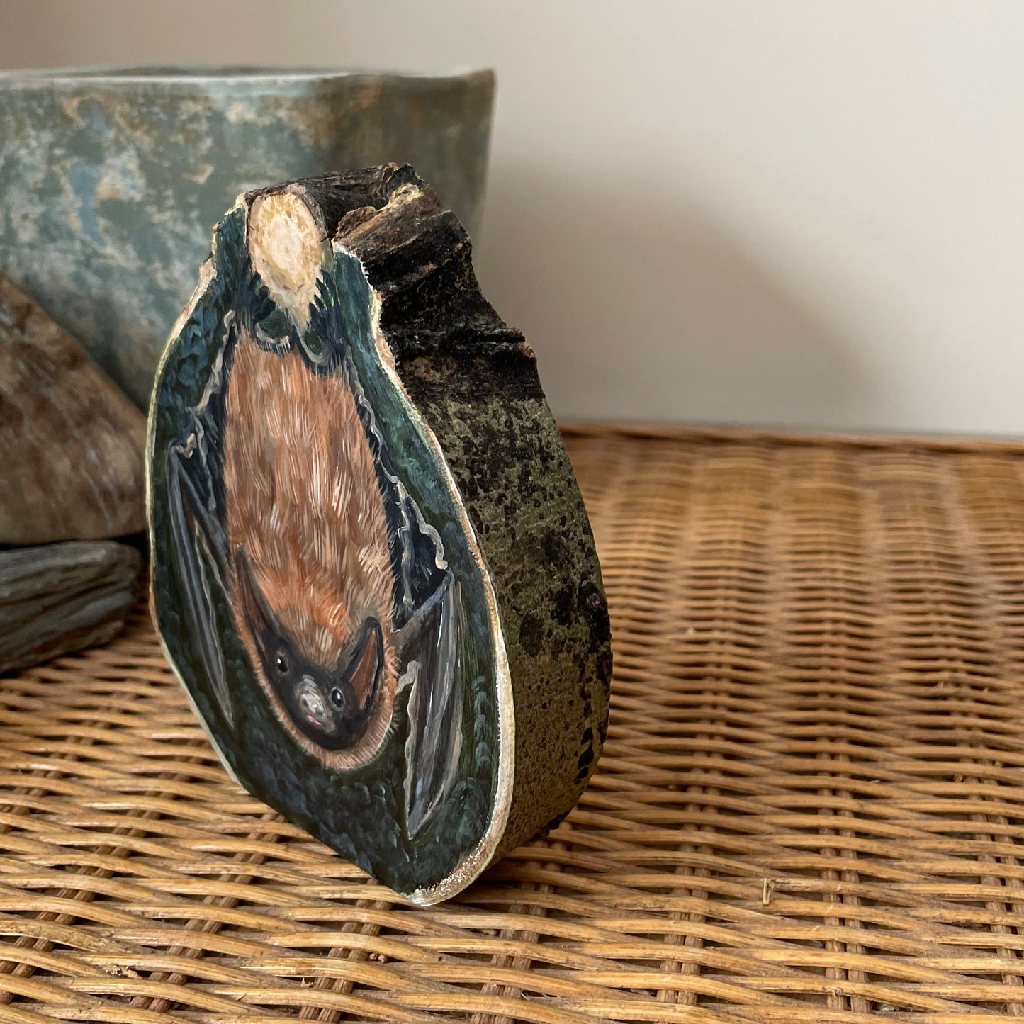 Painting of a Brown bat hanging upside down from a tree knot against a dark forest green background on an aspen wood slice with bark framing the outside